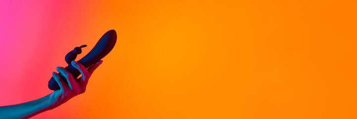 Woman's hand holding vibrator, massager over yellow-pink background in neon light. Concept of toys...