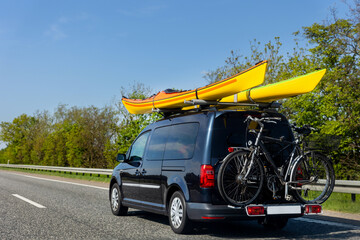 Back view modern black family wagon van car with mounted roof kayak and bike tail carrier driving european highway road against blue sky summer day. Lifestyle travel adventure trip journey concept - Powered by Adobe