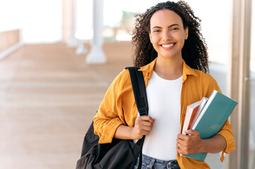 Close-up of a cheerful lovely curly haired brazilian or hispanic female student, with a backpack,...