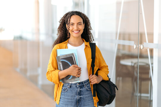 Positive trendy female student. Happy lovely brazilian or hispanic female student, with a backpack, hold books and notebooks in her hand, stand near the university campus, looks and smile at camera