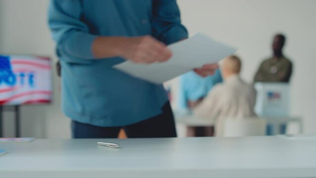 Close-up shot from polling booth at electoral precinct, of multiethnic American citizens in casual clothes queuing to cast their vote, walking up, marking ballot and leaving, USA flag in background