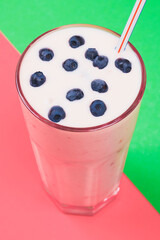 Glass of cold milkshake with fresh blueberries on top. Drink in a glass with a straw on a green and pink background. Multicolored background.
