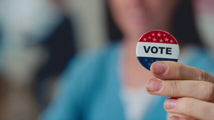 Close-up pan selective focus shot of fingers of unrecognizable blurred Caucasian or Asian woman holding election campaign vote badge, with stars in colours of USA flag