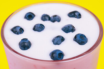 Glass of cold milkshake with fresh blueberries on top. Drink in a glass with a straw on a yellow background.