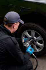 Auto mechanic in a cap changing a wheel from a black car using a drill in a tire shop on the street...