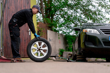 Auto mechanic in a cap changing a wheel from a black car using a drill in a tire shop on the street...