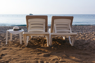 Two sun loungers and a table with a straw hat on a beach. Blue sea background. Silhouette of motorboat on the horizon. Vacatoin 