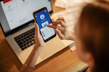 Young woman using a banking app on her smart phone while working on a laptop from home in the kitchen