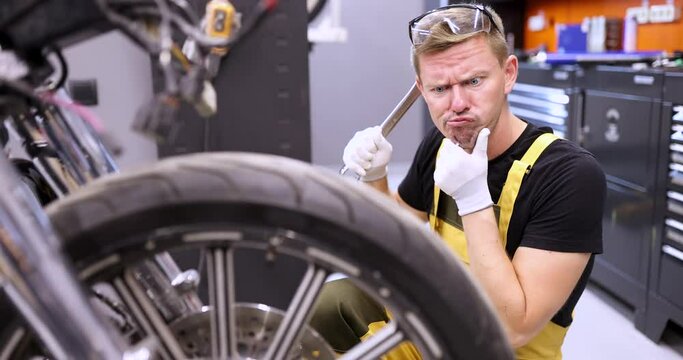Emotionally pensive auto mechanic holds wrench and stares in surprise at motorcycle in service center. Auto transport service concept