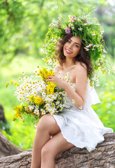 Fototapeta premium Portrait of a cute beautiful girl in a wreath and a white dress, sitting on a big tree in the forest. Ivan Kupala holiday.