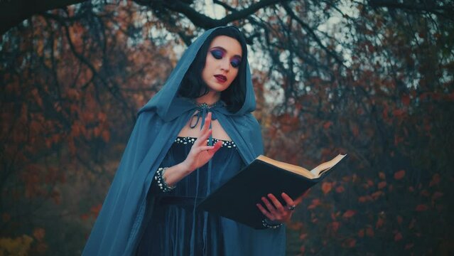 fantasy halloween woman witch creating magic in autumn mystical forest, red orange leaves. Lady witch holds spell book waving hand fages fall. Blue dress silk cape hood old style, girl holiday makeup