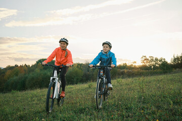 Fototapeta premium Mother and son ride a bike. Happy cute boy in helmet learn to riding a bike in park on green meadow in autumn day at sunset time. Family weekend.