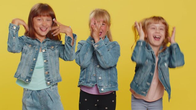 Funny happy teenage girls making playful silly facial expressions and grimacing, fooling around, showing tongue. Little children sisters. Three cute siblings kids isolated on studio yellow background