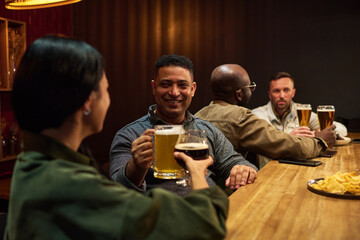 Smiling man with mug of foaming beer toasting with young brunette woman while both standing by wooden bar counter and enjoying party