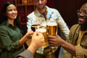 Focus on hands of four young intercultural friends clinking with glasses of fresh cool beer while having fun and enjoying party or gathering in bar