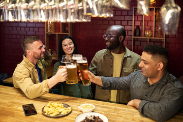 Cheerful intercultural friends with glasses of beer clinking over bar counter with snacks while celebrating victory of their football team at leisure