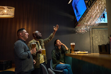 Side view of young intercultural friends having beer and watching hockey broadcast while one of them expressing tension and pointing at screen