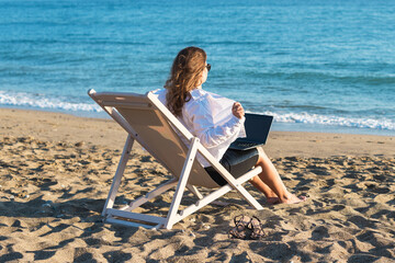 Back view of alone young brunette woman freelancer in office clothes with a laptop looking on the sea sitting on the deck chair on beach. Summer concept