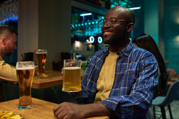 Happy young African American man in blue checkered shirt holding mug of cool foaming beer while sitting by table in pub in front of buddy