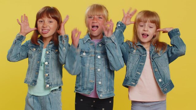 Teenage girls showing tongue making faces at camera, fooling around, joking, aping with silly face, teasing, bullying, abuse. Little children sisters. Three siblings kids isolated on yellow background