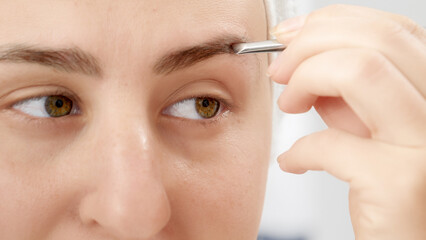 Macro shot of woman plucking eyebrows with small cosmetic tweezers at home. Concept of beautiful female, makeup at home, skin care and domestic beauty industry.