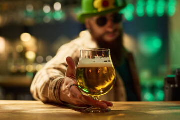 Focus on hand of bearded barman in green hat and checkered shirt putting glass of cool foaming beer on wooden counter in pub