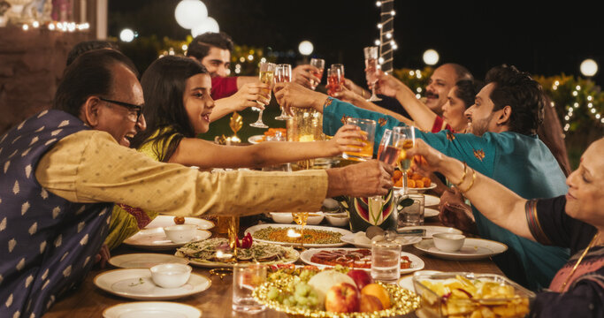 Happy Indian Family Having A Feast And Celebrating Diwali Together: Group Of People Of Different Ages In Their Traditional Clothes Raising Glasses And Making A Toast In A Backyard Garden