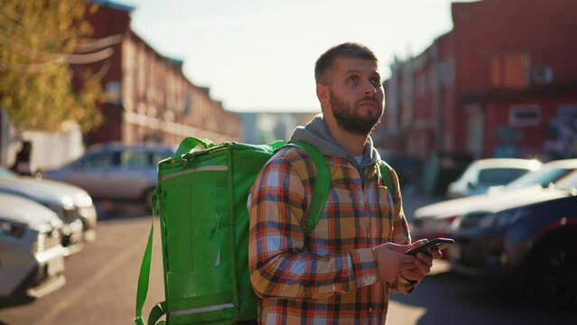 Man courier finding address on map in smartphone on city street looking around. Food delivery guy navigates in phone application with green huge backpack on spine. Fast delivery, courier work concept.