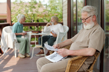 Bearded senior man with grey hair sitting in wicker armchair on patio of retirement home and reading newspaper with latest news - Powered by Adobe