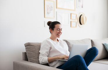 Fototapeta premium Young woman working on laptop. Millennial girl working remote job from home, sitting on the cozy sofa, smiling. 