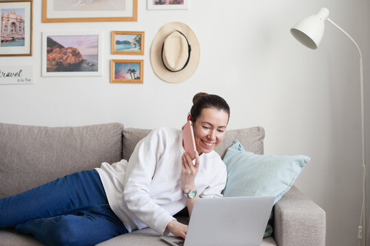 Young woman working on laptop. Millennial girl working remote job from home, using mobile phone, sitting on the cozy sofa, smiling. 