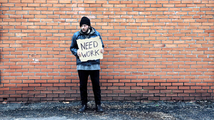 Homeless holds piece of cardboard with inscription Need Work. Refugee is at brick wall looking for...