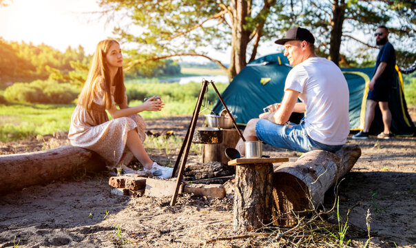 A guy and a young girl sit on a log in nature and talk against the backdrop of scum and put up a tourist tent. The concept of summer recreation in nature on the sandy bank of the river, picnic