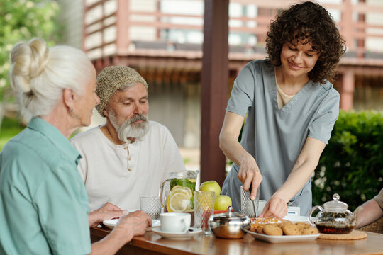 Young pretty caregiver cutting cake or pie for group of senior people sitting by table served for breakfast and waiting for tea with dessert