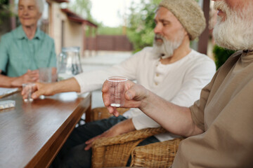 Focus on hand of senior man holding transparent cup with pills while sitting by wooden table in front of camera against other elderly people