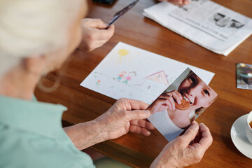 Hands of elderly woman sitting by wooden table in retirement home and holding photo of her cute granddaughter biting homemade oat cookie