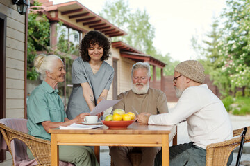 Young smiling carer in uniform and senior female patient of retirement home looking at paper document next to elderly bearded men