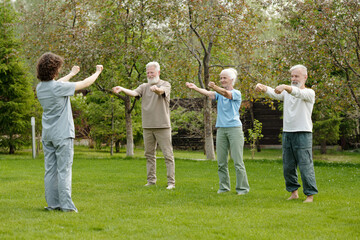 Fototapeta premium Row of senior people in activewear repeating after their instructor or caregiver while doing physical exercises on green lawn in the garden