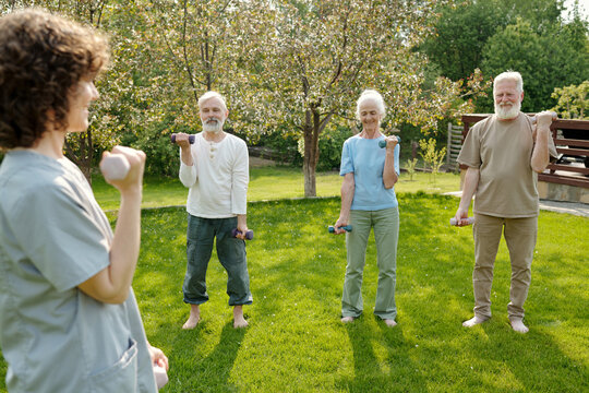 Group of active senior people doing exercise with dumbbells and looking at instructor or nurse in uniform during course of rehabilitation therapy