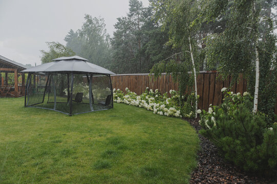 Summer heavy rain at the cottage, yard with green lawn and flowers, picnic gazebo