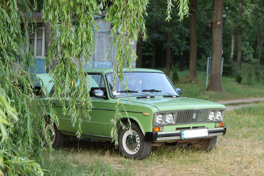 A Soviet green VAZ 2106 car with an open trunk parked next to a willow tree on the beach.