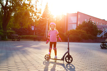 A little caucasian girl in a pink protective gear laughs and grimaces near a scooter against the backdrop of a summer sunset. © HENADZY