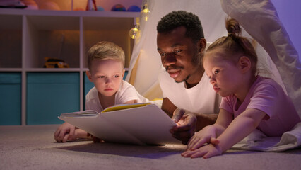 African American uncle reads bedtime stories to kids siblings in cozy play tent.
