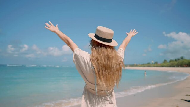 Excited woman tourist enjoying resting on sandy tropical beach rising hands up touching warm air, rear view. Blonde female standing on shore admiring turquoise ocean water. Travel, tourism, vacation.