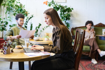 Young serious female freelancer or solopreneur sitting in cafe in front of laptop, typing an looking at screen while watching online video