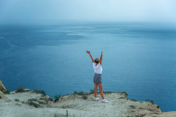 Heavy rain on the sea on a sunny day. A young woman in a white T-shirt stands on the edge of a high cliff. Magnificent landscape.