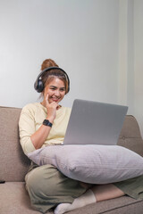 Young beautiful asian woman relaxing and listening to music using headphones, she is lying in sofa. Young woman enjoying at home using laptop.