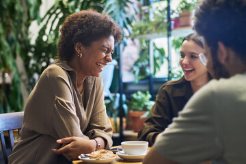 Young laughing African American woman sitting by table with coffee and snack in front of her friends while enjoying pastime with them in cafe