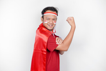 Excited Asian man wearing a red top, flag cape and headband, showing strong gesture by lifting his...