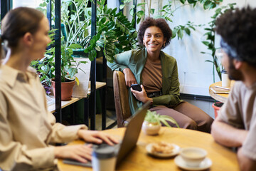 Focus on young smiling African American businesswoman with smartphone looking at female colleague...